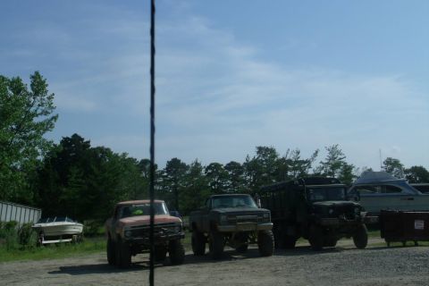 1978 Ford Bronco - Several modified trucks are parked outdoors on a sunny day, with trees and a boat in the background.