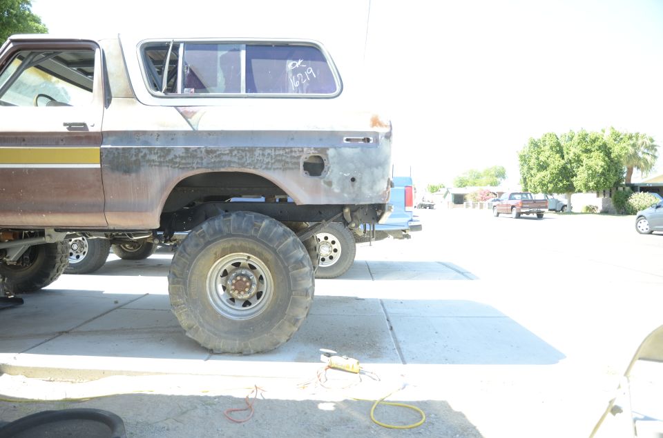 Side view of a lifted 1978 Ford Bronco with large tires, showing rust and missing rear bumper. Another vehicle is parked behind it