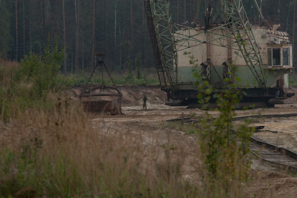 Giant excavator machine with a dragline bucket hanging nearby, a person stands between them in a dirt quarry