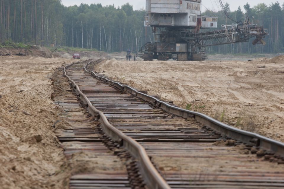 Railroad tracks curve through a sandy area towards a large excavator; a red vehicle is visible in the distance