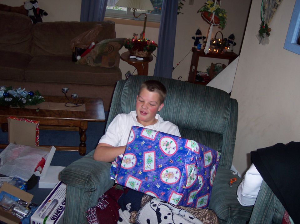 Boy in armchair opening a large blue gift with Christmas patterns. Presents and wrapping paper are scattered around him