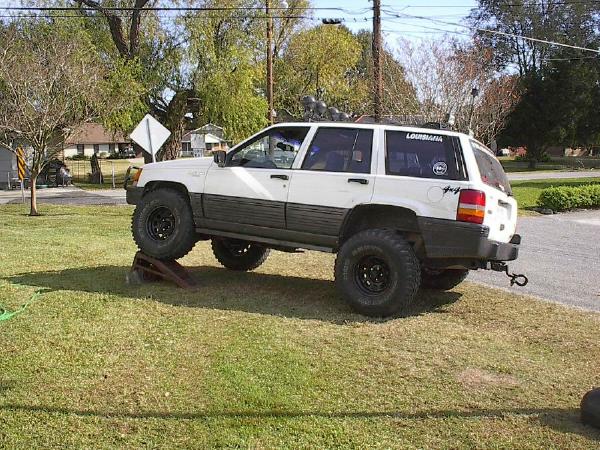 Lifted white Jeep Grand Cherokee on off-road tires, front wheel on a ramp in a grassy yard, side view
