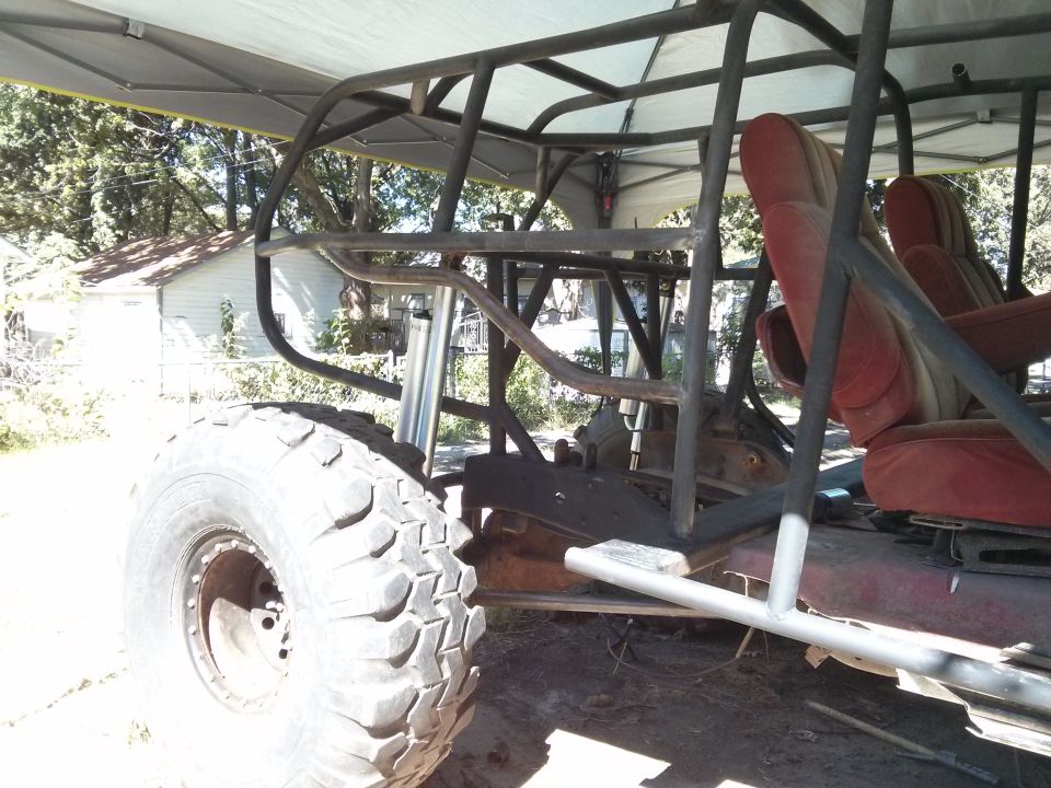 Custom buggy frame with red seats and large off-road tire, parked under a canopy