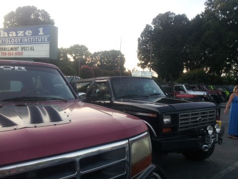 1986 Ford Bronco - A black SUV with a custom front bumper and auxiliary lights is parked in front of a sign for "Haze 1 Cosmetology Institute."