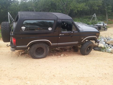 1986 Ford Bronco - This black SUV is parked on a dirt lot with trees in the background and another vehicle on a trailer behind it.