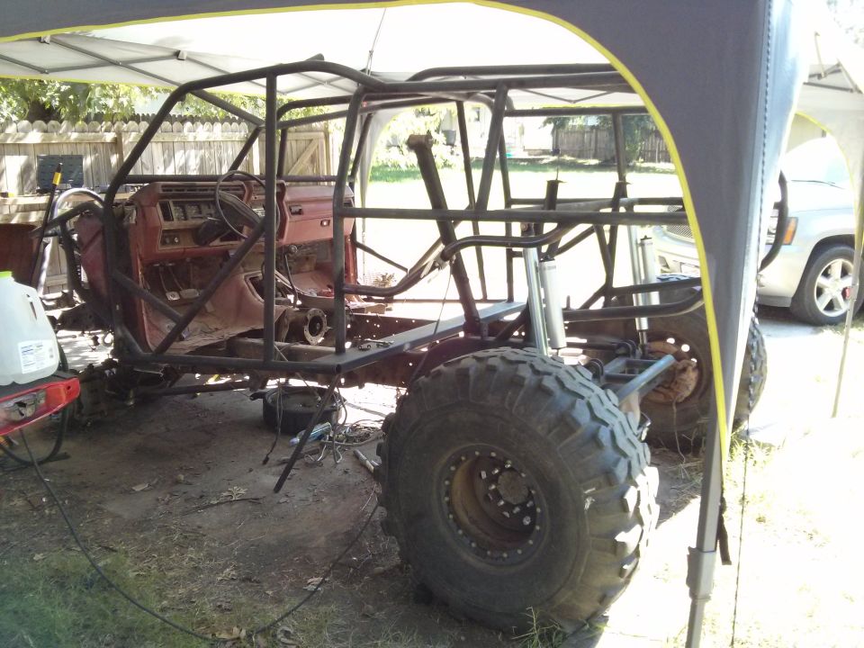 Off-road vehicle chassis with roll cage, red interior, large tires, and a white SUV in the background