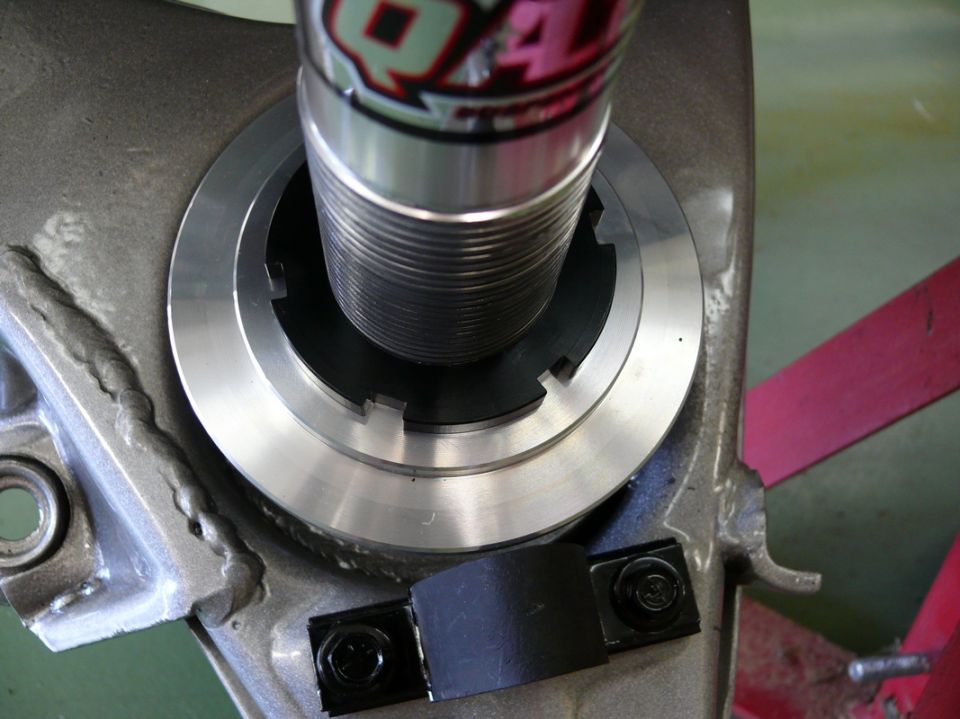 Close-up of a shock absorber assembly with a threaded adjustment collar and a black rubber strap secured by two bolts