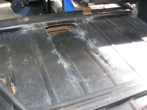 Close-up of a vehicle's metal floor pan with visible welding repairs and a cut-out section