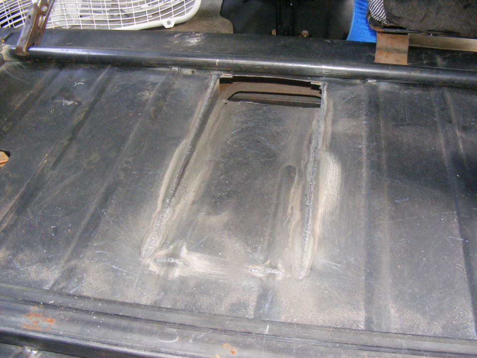 Black metal floor pan with a rectangular opening and visible weld repairs