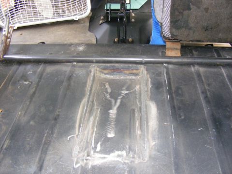 Close-up of a vehicle's metal floor with a welded repair patch. A seat and a fan are visible in the background