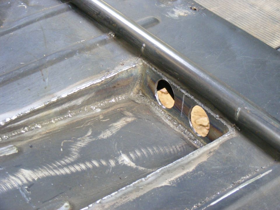 Close-up of metal frame with two circular cutouts and a welded bar. Some weld marks and scratches visible