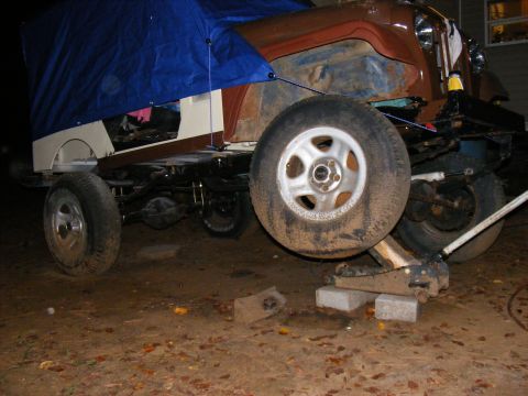 Off-road vehicle chassis on jack, partially covered by blue tarp. Brown body panels visible