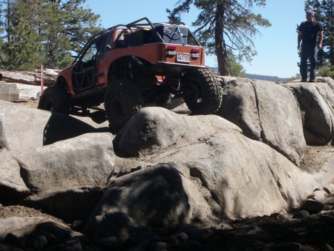 1961 International Scout 800 - An orange off-road vehicle navigates a rocky terrain with a person standing on a rock in the background.
