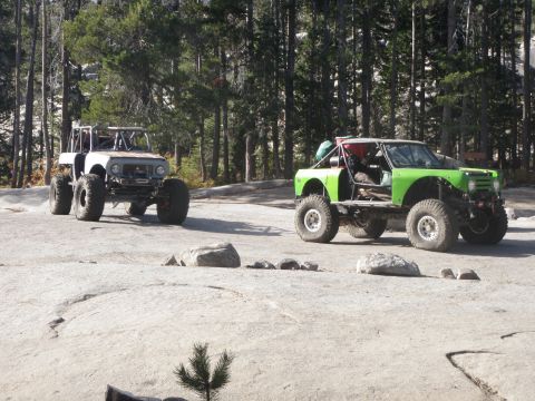 1961 International Scout 800 - Two heavily modified off-road vehicles are parked in a natural, rocky environment with trees in the background.