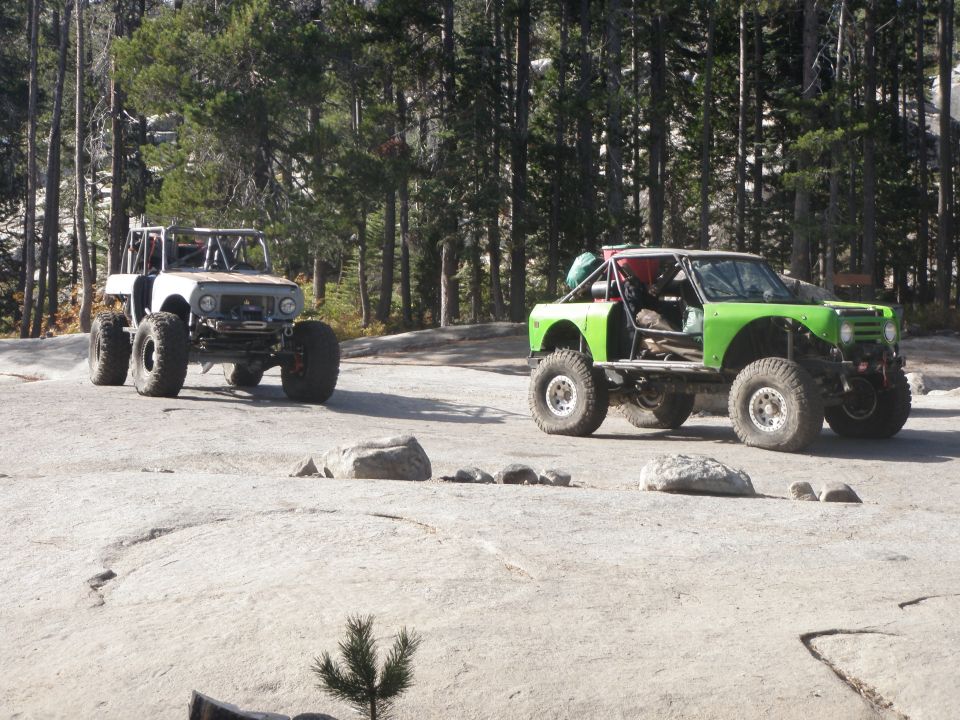 Two custom off-road vehicles parked on a rocky, wooded area. One is white, the other is green