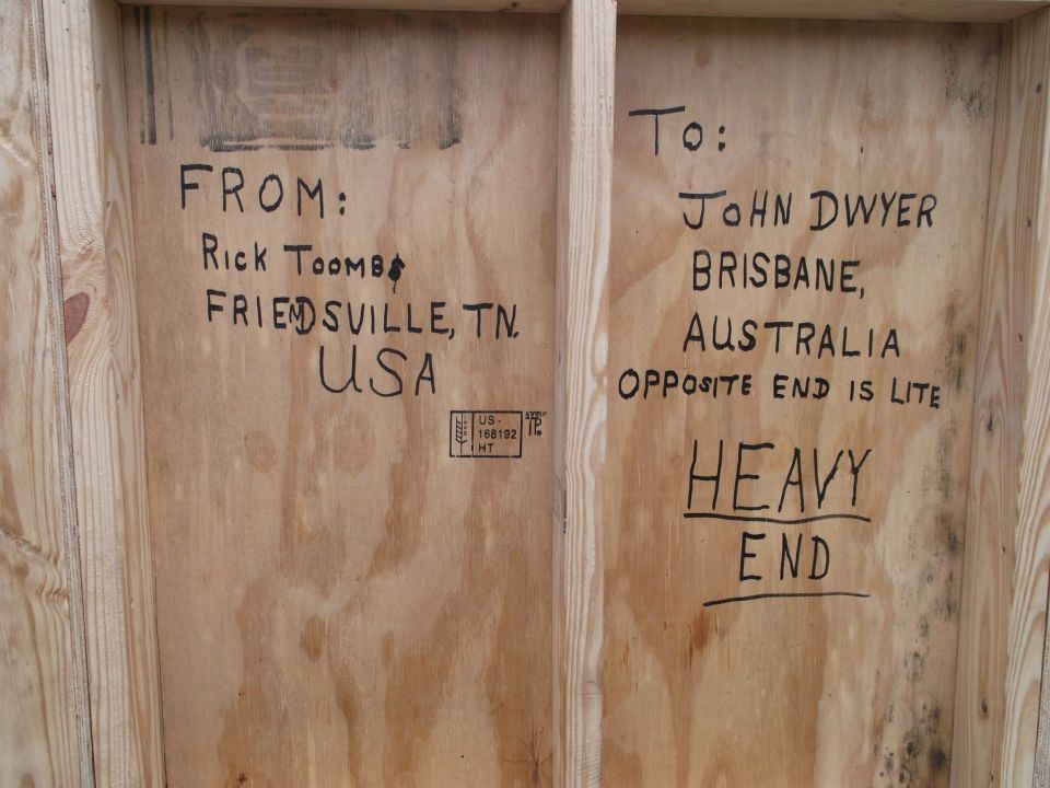 Wooden crate with handwritten shipping information: FROM: Rick Toombs, Friendsville, TN, USA. TO: John Dwyer, Brisbane, Australia. Opposite end is lite. Heavy end