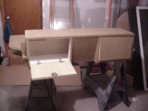 1993 Ford Bronco - Carpet-wrapped custom storage console on sawhorses in a basement, with one hinged plywood door dropped open on chrome hinges and a chest-st…
