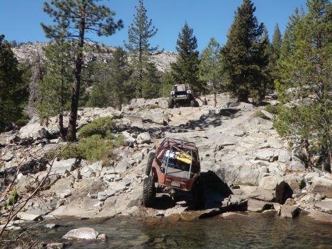 1961 International Scout 800 - Two modified off-road vehicles traverse a challenging rocky path with trees and a clear stream.