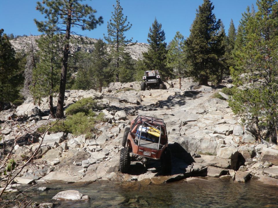 Two off-road vehicles navigate a rocky, wooded trail near a stream