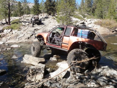 1961 International Scout 800 - Two heavily modified off-road vehicles traverse a rocky stream bed in a mountainous, wooded area.