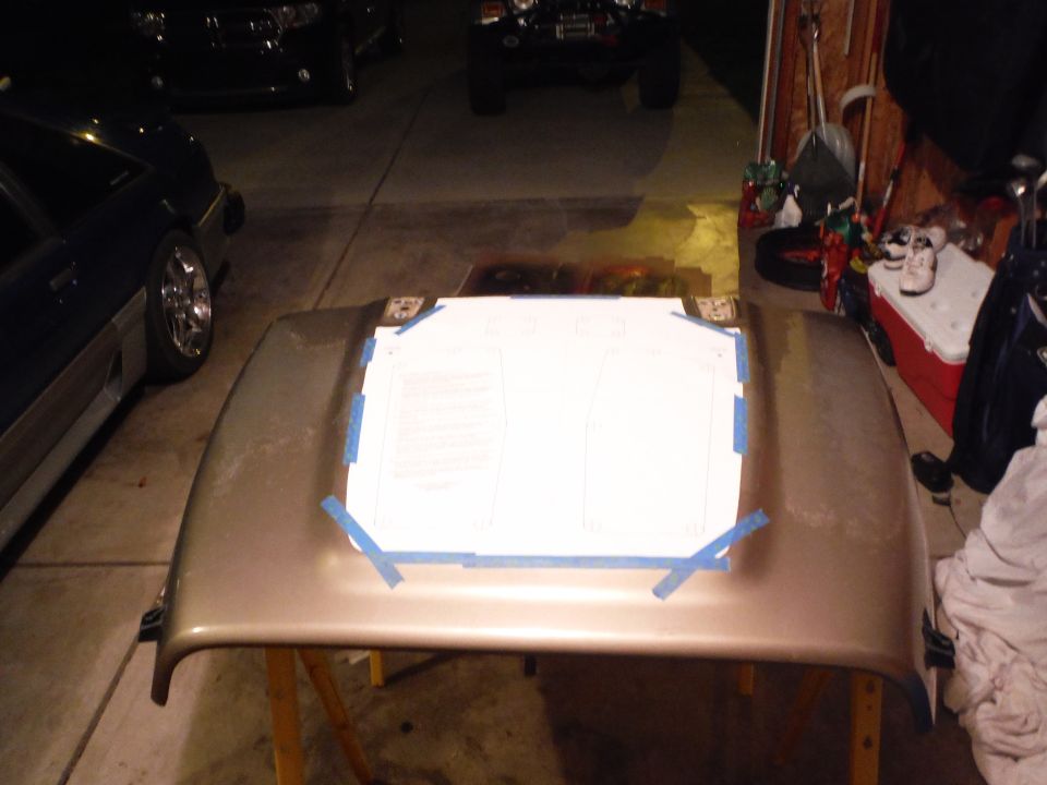 Car hood with a paper template taped to it, parked in a garage with other vehicles visible