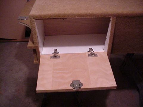 1993 Ford Bronco - Detail of a carpeted custom console compartment with its plywood drop-down door open, held by two chrome cabinet hinges and closed by a cen…