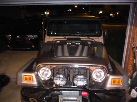 2005 Jeep Wrangler - A Jeep Wrangler is parked in a garage with a black SUV behind it. The Jeep has an aftermarket hood scoop and PIAA lights.