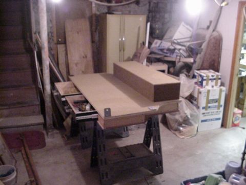 1993 Ford Bronco - Wide basement-workshop view of a partly built particle-board cargo console resting on folding sawhorses, its lower drawer pulled open to re…