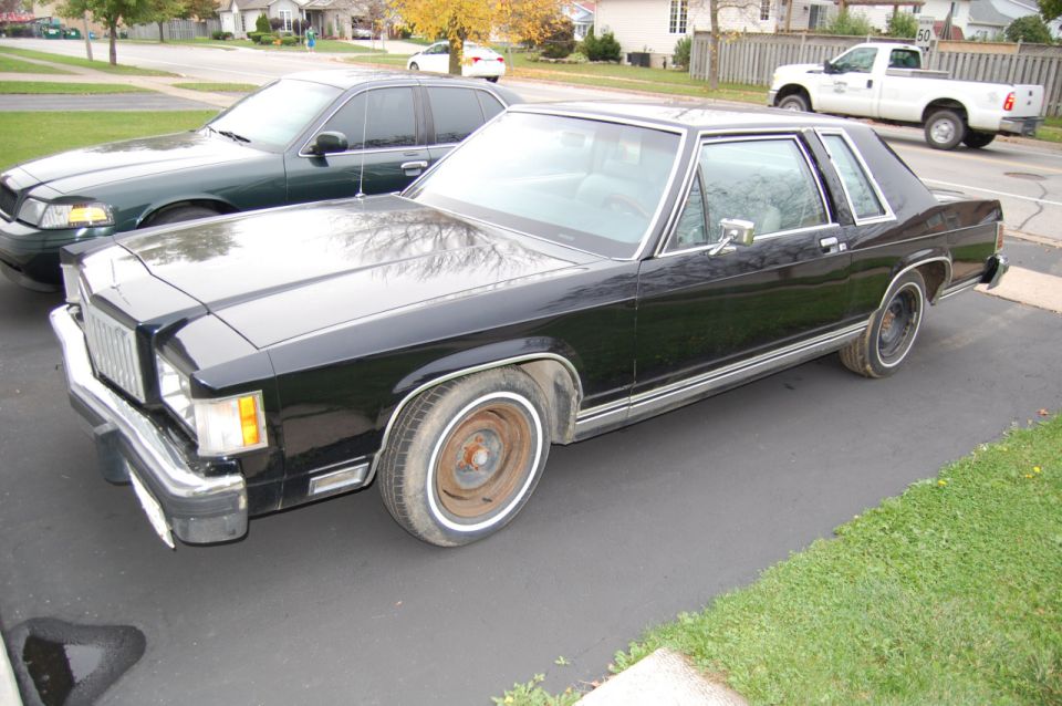 Black 1985 Mercury Grand Marquis parked next to a dark green sedan. White pickup truck in background