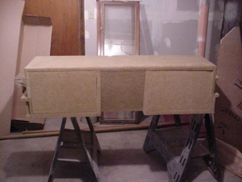 1993 Ford Bronco - Custom cargo console fully wrapped in tan carpet, sitting on black sawhorses in a basement and viewed straight on from the front. Two flush…