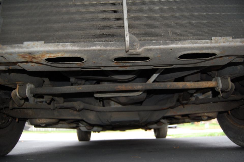 Undercarriage of a vehicle showing the radiator, suspension components, and exhaust system. Some rust is visible on the frame