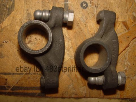 1969 Ford Fairlane - Close-up of two engine rocker arms, showing their metallic texture and bolted components against a wood grain background.