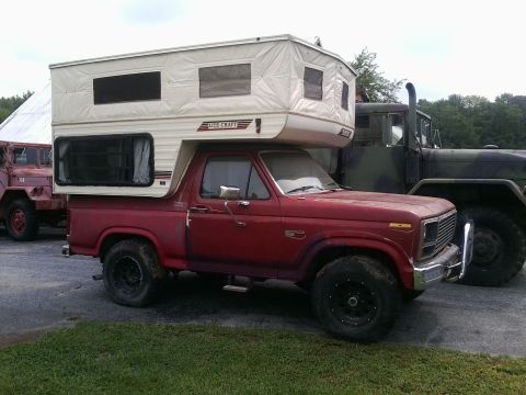 1984 Ford Bronco - A red Ford Bronco with a white camper shell is parked next to a military truck.