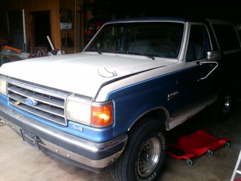 1984 Ford Bronco - A blue and white Ford Bronco is parked in a garage, showing its front and side profile.