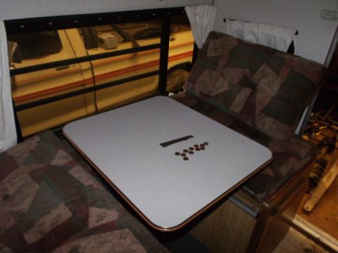 1984 Ford Bronco - View from inside a camper, showing a table with coins and a ruler, with a yellow vehicle seen through the window.