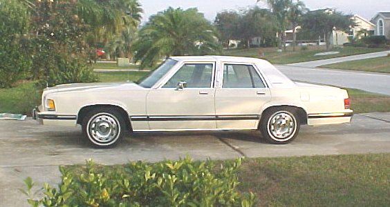 White Mercury Grand Marquis sedan in profile on a driveway with palm trees behind