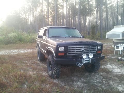 1986 Ford Bronco - A dark colored, lifted 1980s Ford Bronco is parked on a dirt lot with trees in the background and a pop-up camper to the right.