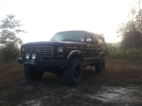 1986 Ford Bronco - A black Ford Bronco is parked in a grassy, wooded area with trees in the background. The SUV features a chrome grille, aftermarket bumper w…