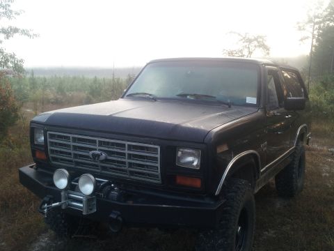 1986 Ford Bronco - A dark-colored, lifted Ford Bronco with a winch and auxiliary lights mounted on the front bumper sits in a misty, outdoor setting.