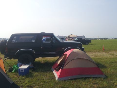 1986 Ford Bronco - A black Ford Bronco is parked in a grassy field next to a red and tan tent. Other vehicles and tents are visible in the background.