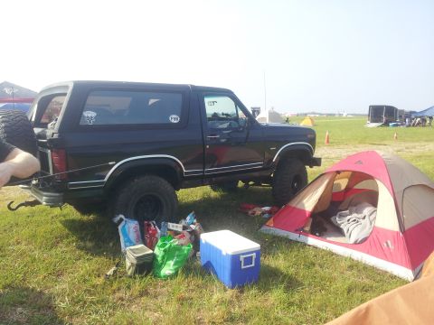 1986 Ford Bronco - A black Ford Bronco is parked on a grassy field next to a red and tan tent. Various camping supplies are scattered on the ground near the v…