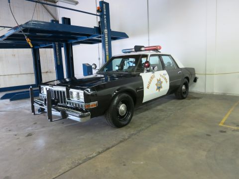 1991 Ford LTD Crown Victoria - black-and-white California Highway Patrol sedan with a roof-mounted light bar sits inside a workshop beside a hydraulic two-post lift.