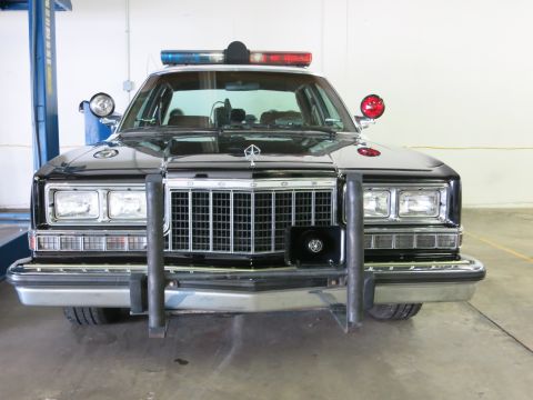1991 Ford LTD Crown Victoria - Front view of a black police car showing a chrome push bumper, roof light bar, red spotlight and a pentastar emblem above the grille.
