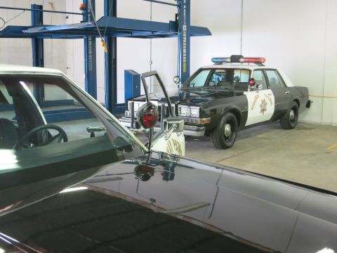 1991 Ford LTD Crown Victoria - black-and-white highway patrol sedan parked under shop lights, viewed across the hood of a dark car with a chrome mirror in the foreground.
