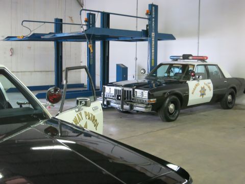 1991 Ford LTD Crown Victoria - black-and-white highway patrol sedan sits beside a two-post lift, with a Highway Patrol door decal visible on a car in the foreground.