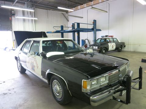 1991 Ford LTD Crown Victoria - black-and-white Ford highway patrol sedan with its hood up and a front push bar, parked in a shop next to another patrol car.