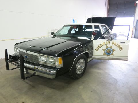 1991 Ford LTD Crown Victoria - black-and-white Ford highway patrol sedan with a push bar and roof light bar, its rear door open showing a gold Highway Patrol star decal.