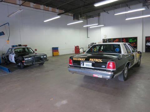 1991 Ford LTD Crown Victoria - Rear three-quarter view of a dark Ford sedan lettered HIGHWAY PATROL on the trunk, parked in a shop beside a black-and-white patrol car.