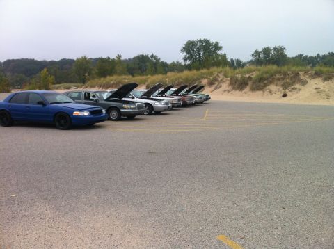 2003 Ford Crown Victoria - A line of sedans, mostly with hoods open, are parked in a lot next to a sandy embankment and trees. The closest vehicle is a blue sedan wit…
