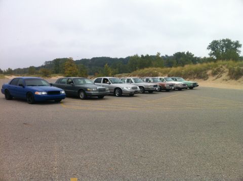2003 Ford Crown Victoria - A line of sedans are parked in a lot with a sandy embankment and trees in the background. The closest vehicle is a blue sedan.